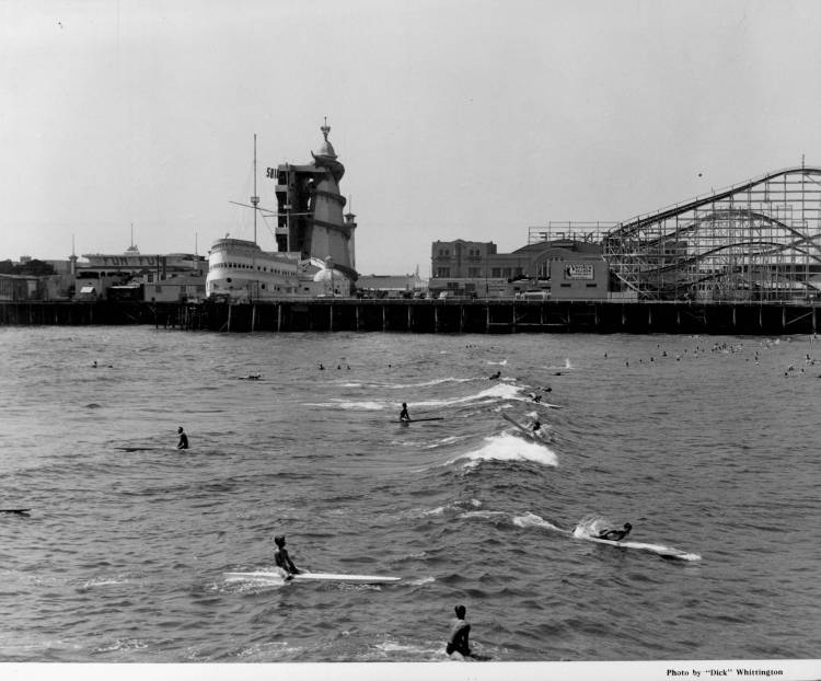1939 - People ride surf boards and paddle boards in front of an amusement park